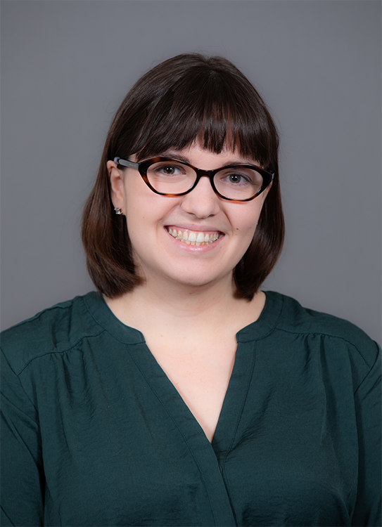 Head shot of a caucasian woman wearing brown oval eyeglasses, with brown shoulder length hair and is wearing a green v-neck shirt, with a blue-grey background.